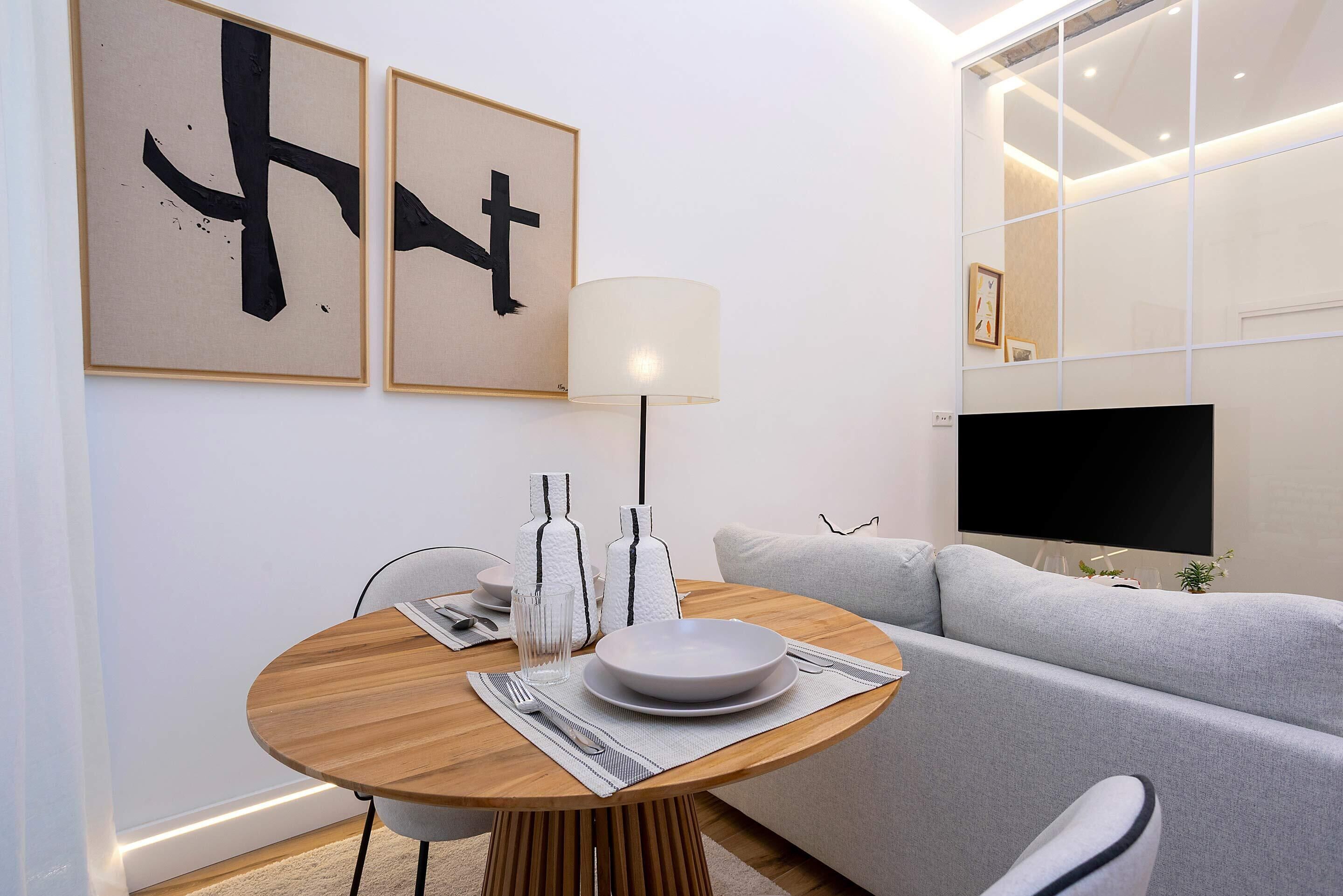 A modern living room with a round wooden dining table, a grey sofa, and a flat-screen TV.