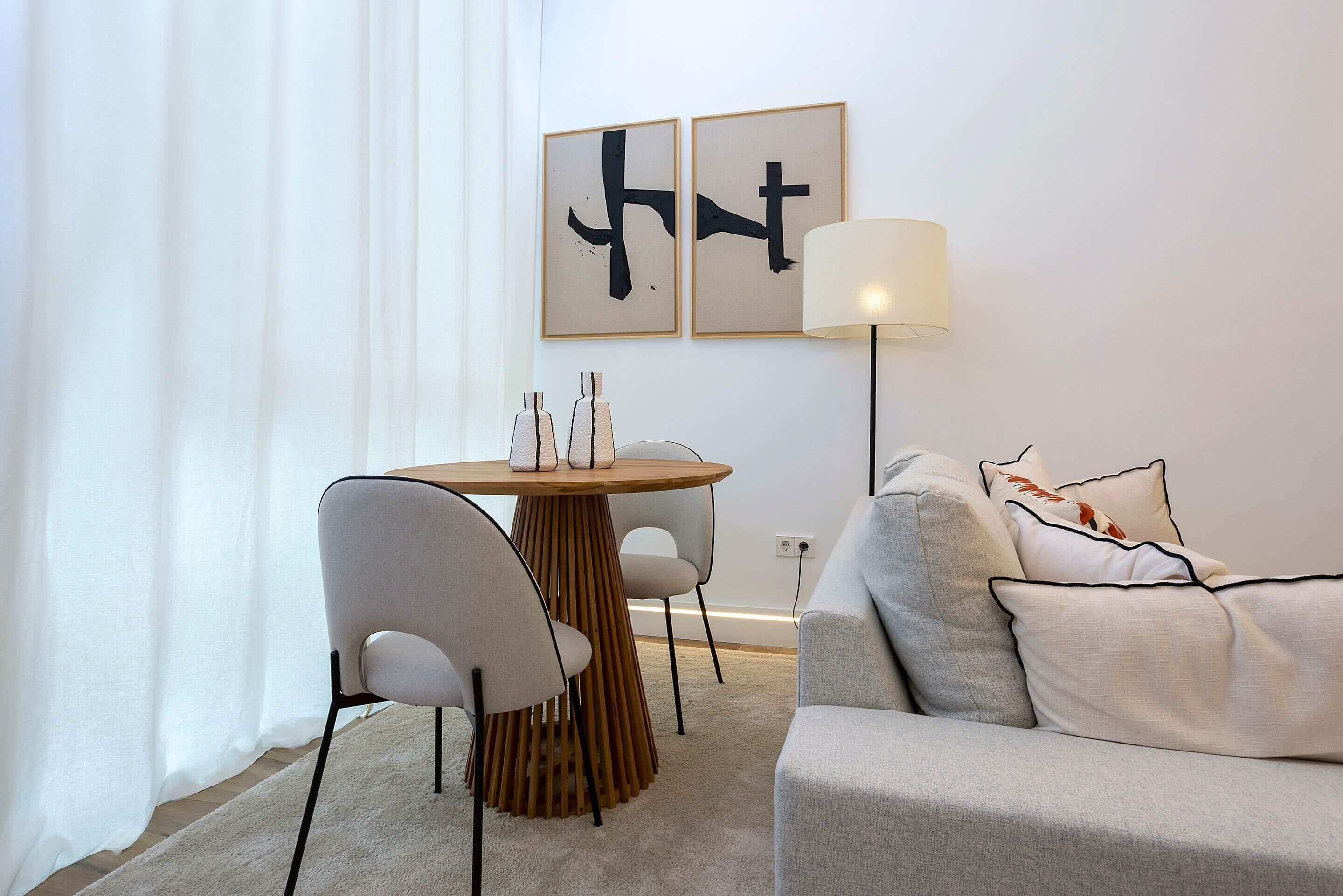 A modern living room with a round wooden table, two grey chairs, a grey sofa with patterned cushions, and two abstract paintings on the wall.