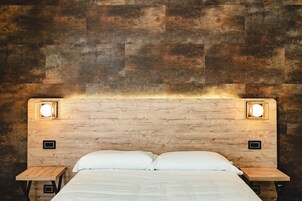 Una cama con ropa blanca, dos mesitas de noche y un cabecero de madera con dos luces fijadas a la pared.