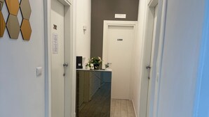 A hallway with light-colored walls, a dark ceiling, and a tiled floor. There are three doors on the left, each with a number plaque. A small table with a vase of flowers is in the center. - LA PIAZZA SUITE (Naples)