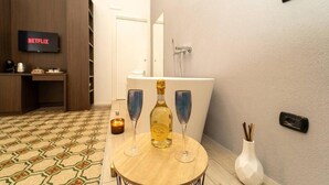 A room with a patterned floor, a table with a bottle and glasses, a TV displaying 'NETFLIX', a bathtub, and a wall-mounted light switch. - LA PIAZZA SUITE (Naples)