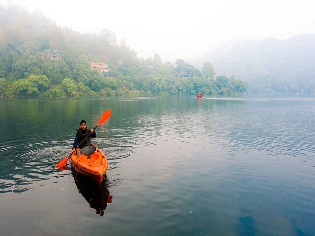 Una persona practica kayak en un lago tranquilo, con una ladera boscosa al fondo.