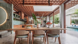 A spacious dining area with a wooden table and woven chairs, a kitchen with a modern design, and a view of a swimming pool. - Vinaka Villa By Betterplace (Canggu)