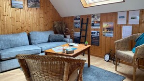 Une pièce chaleureuse avec un plafond en pente, des murs en bois, un canapé bleu, des fauteuils en osier, une petite table avec des papiers et un vélo.
