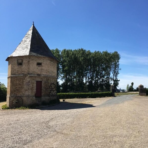 A small stone building with a conical roof, a red door, and a large wheel on the ground. - Chambres d hotes à la ferme  (Forest-Montiers)