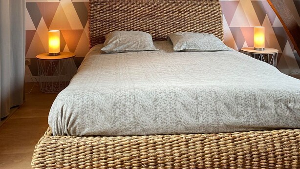 Une chambre à coucher avec un mur orné d’un motif géométrique, une tête de lit en bois, un cadre de lit en osier et deux lampes de chevet.