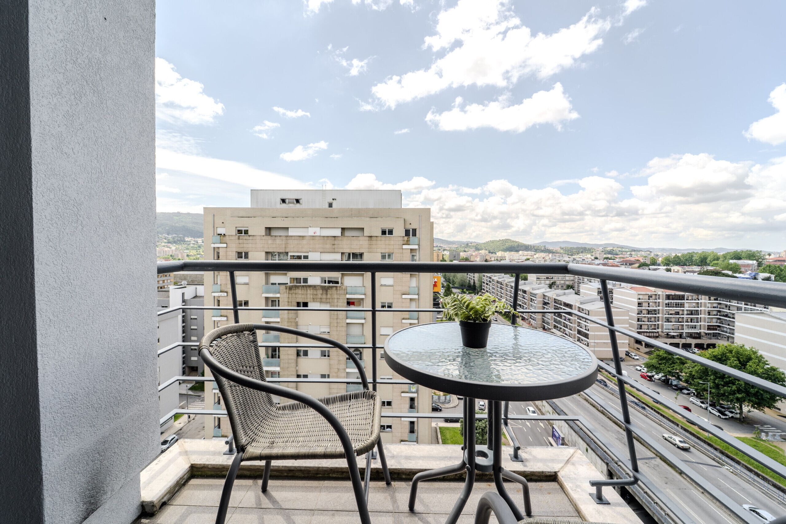 Ein Balkon mit rundem Tisch und zwei Korbstühlen, mit Blick auf eine Stadtlandschaft mit Gebäuden und einer Straße.