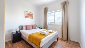 Un dormitorio ordenado con cama, mesita de noche, ventana con cortinas y vista a la ciudad.