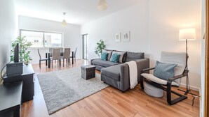 Una sala de estar moderna con un sofá gris, una silla negra, un comedor con sillas y una mesa, y un ventanal que proporciona luz natural.