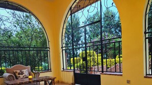 A spacious room with large arched windows, a wooden table, a chair, and a black pot with red flowers. - A  farmhouse on the Tea farm estate (Kiambu County)