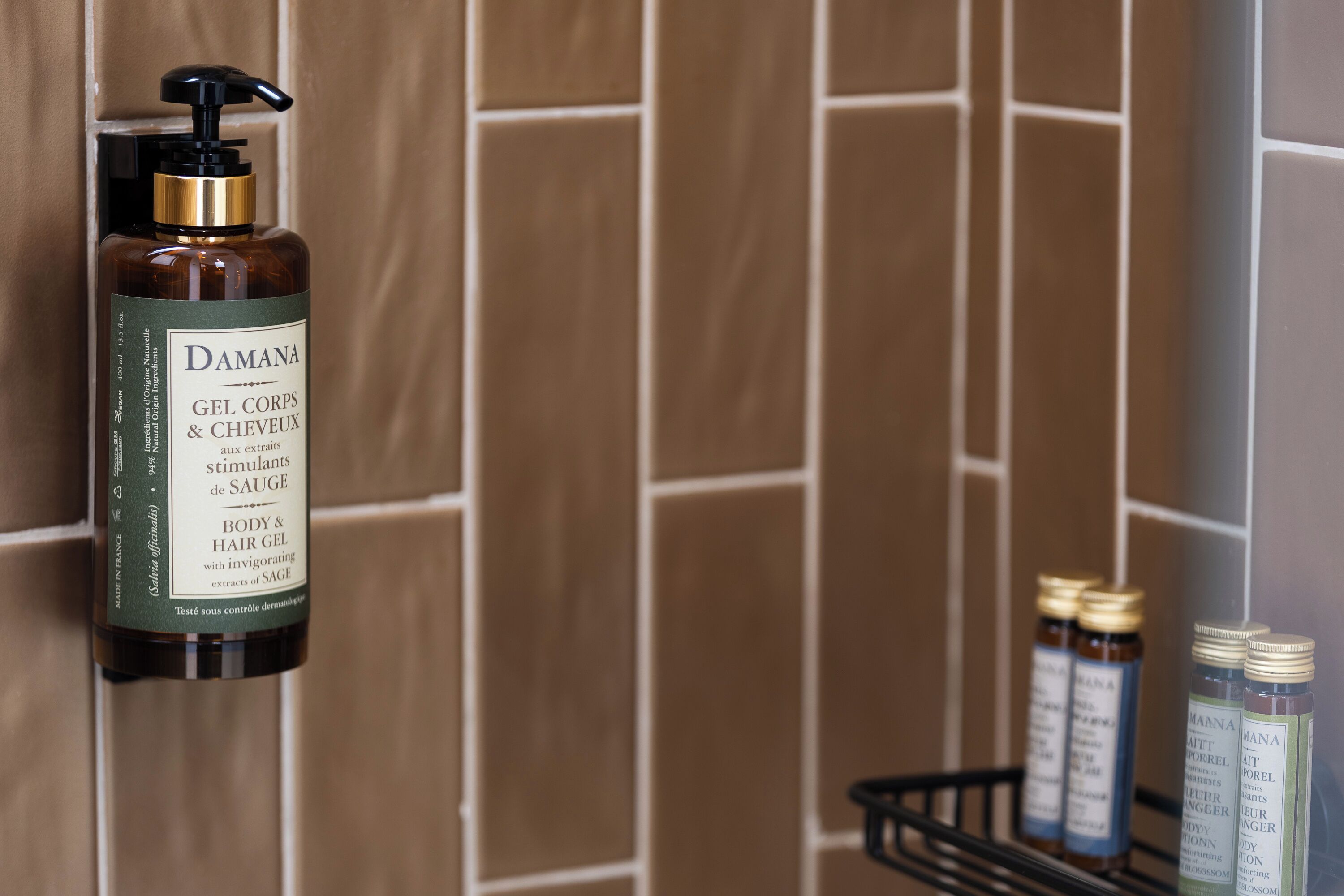 A bathroom with a wall-mounted bottle of Damana body and hair gel, and several smaller bottles on a shelf.
