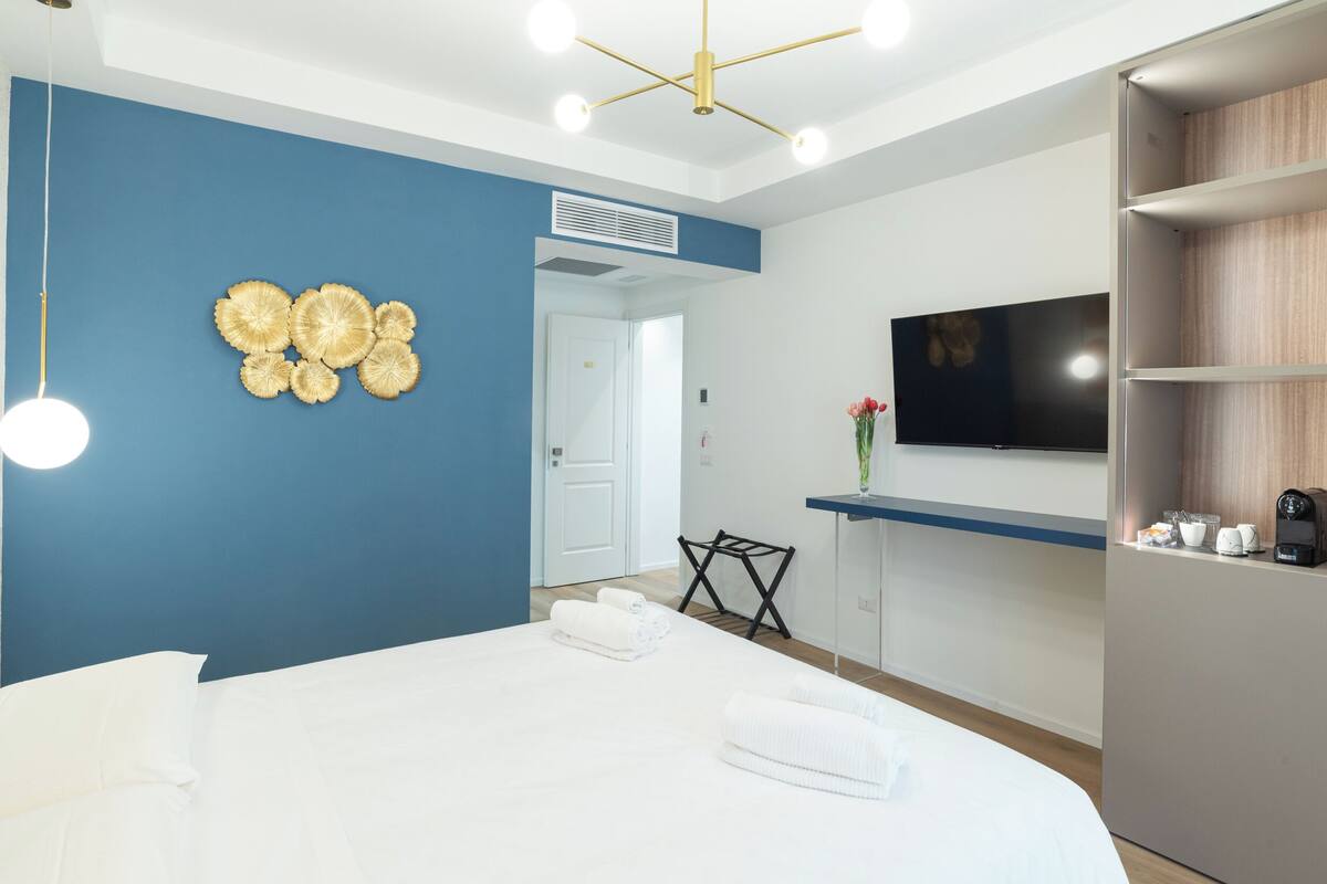 Hotellihuone, jossa on suuri sänky, taulutelevisio, työpöytä tuolilla sekä seinälle kiinnitetty hylly, jossa on hygieniatarvikkeita.