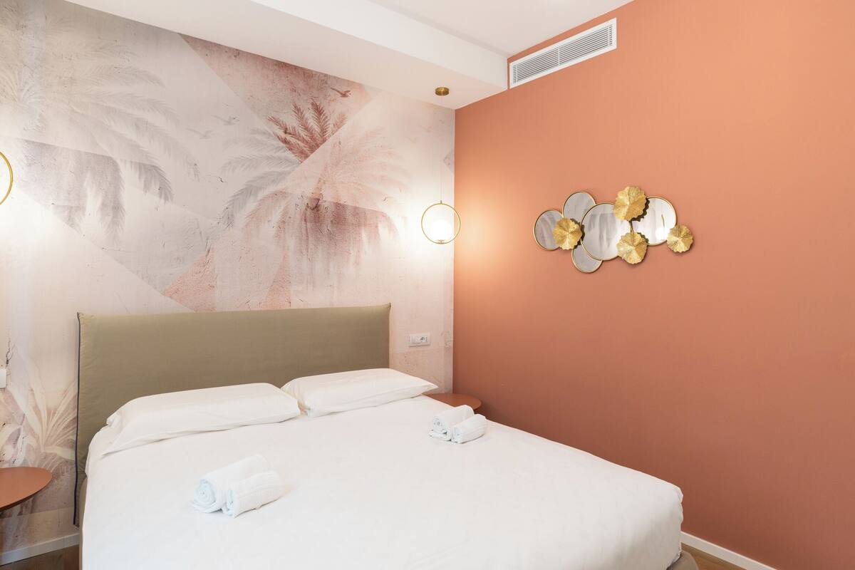 Hotellihuone, jossa on sänky, seinä, jossa on palmunlehtiä kuvaava seinämaalaus, pyöreä peili, jossa on kultaisia yksityiskohtia, ja pieni pöytä, jossa on lamppu.