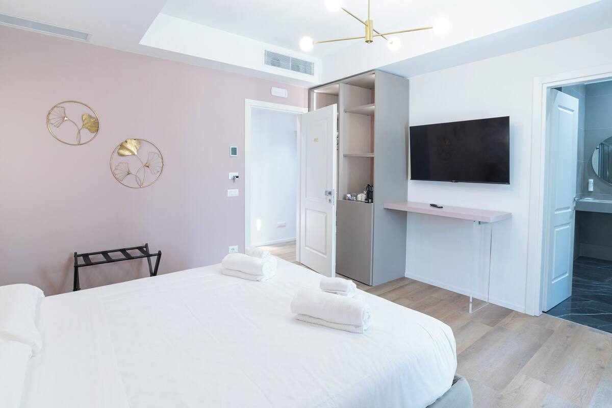 Moderni hotellihuone, jossa on sänky, televisio, vaatekaappi ja kylpyhuone, joka näkyy avoimesta ovesta.
