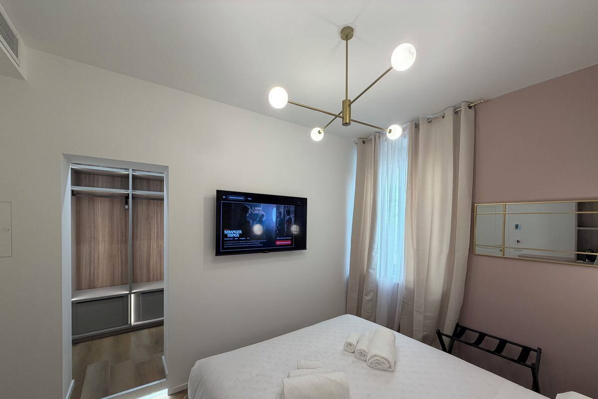 Hotellihuone, jossa on sänky, seinälle asennettu televisio, vaatekaappi ja ikkuna, jossa on verhot.