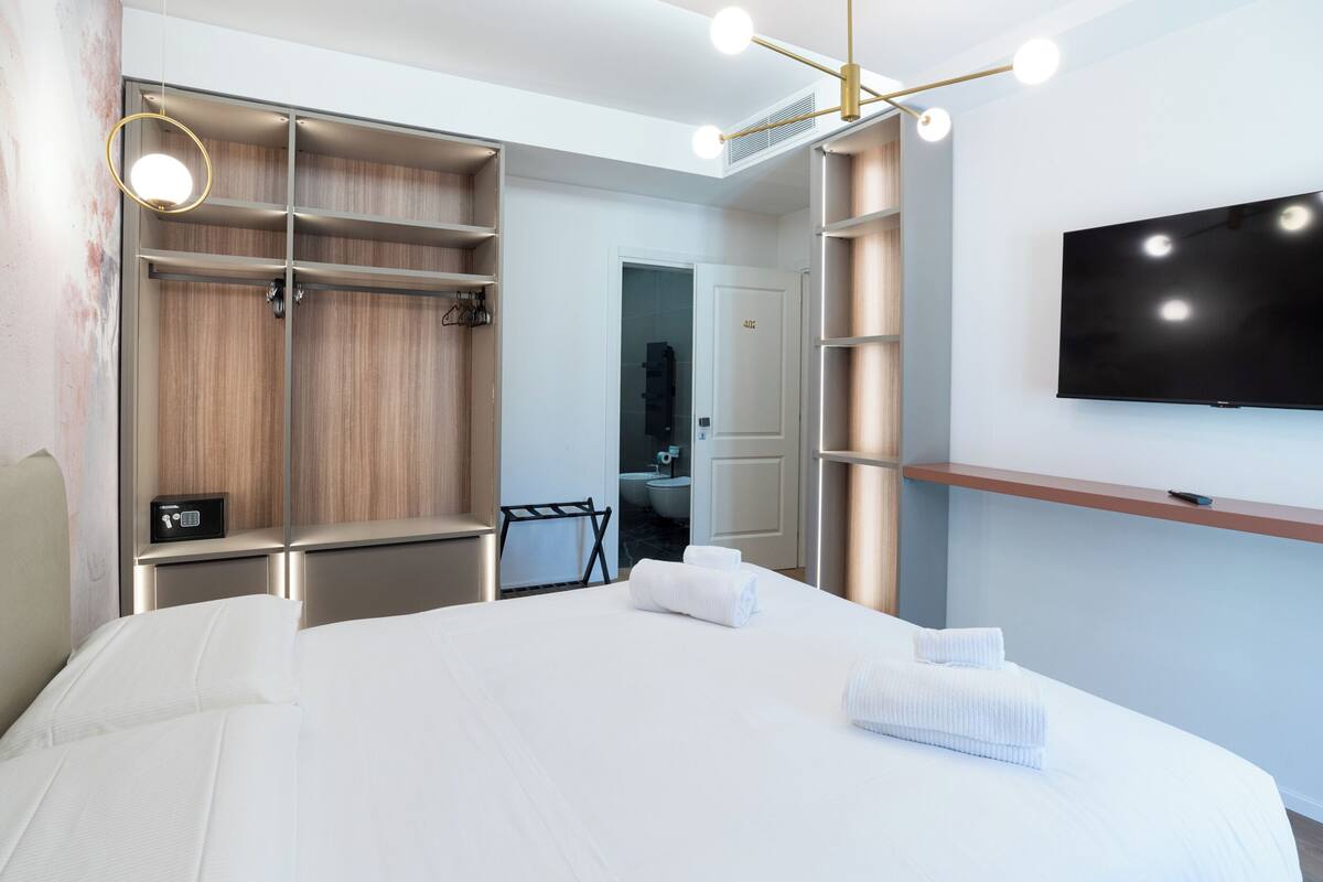 Hotellihuone, jossa on suuri sänky, seinälle asennettu televisio, erillinen vaatehuone ja kylpyhuone, joka näkyy avoimesta ovesta.