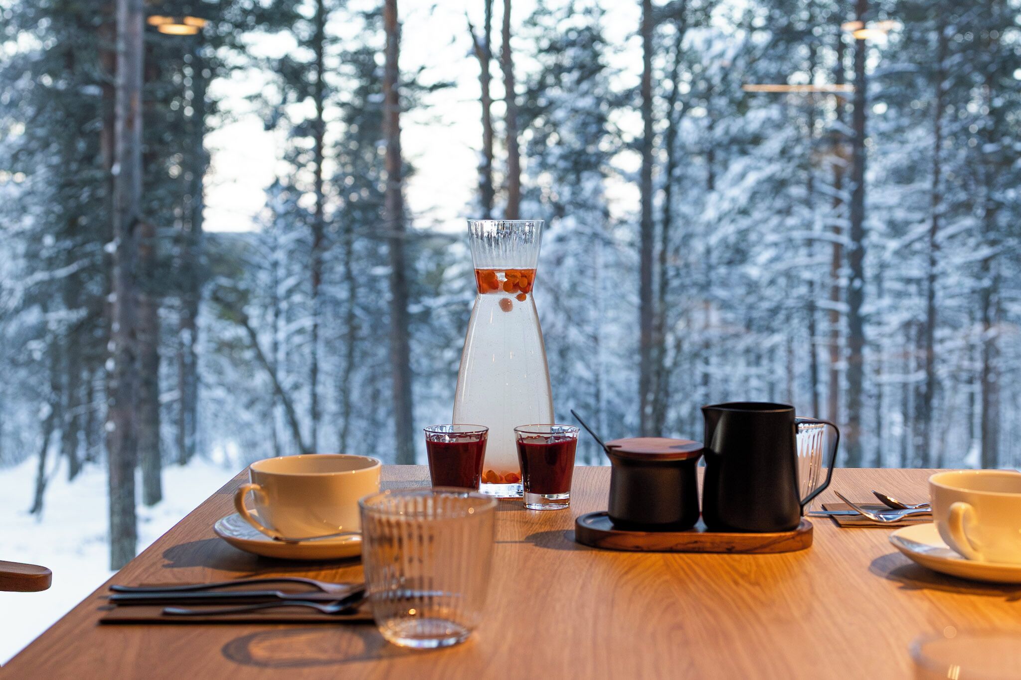 Une table en bois, agrémentée d’un service à café comprenant une carafe en verre, des tasses et une bouilloire, le tout placé devant un paysage de forêt enneigée.