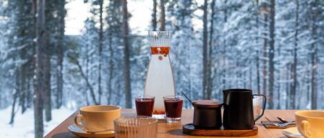 Una mesa de madera con una disposición para café, que incluye una jarra de vidrio, tazas y una tetera, frente a un paisaje de bosque nevado.