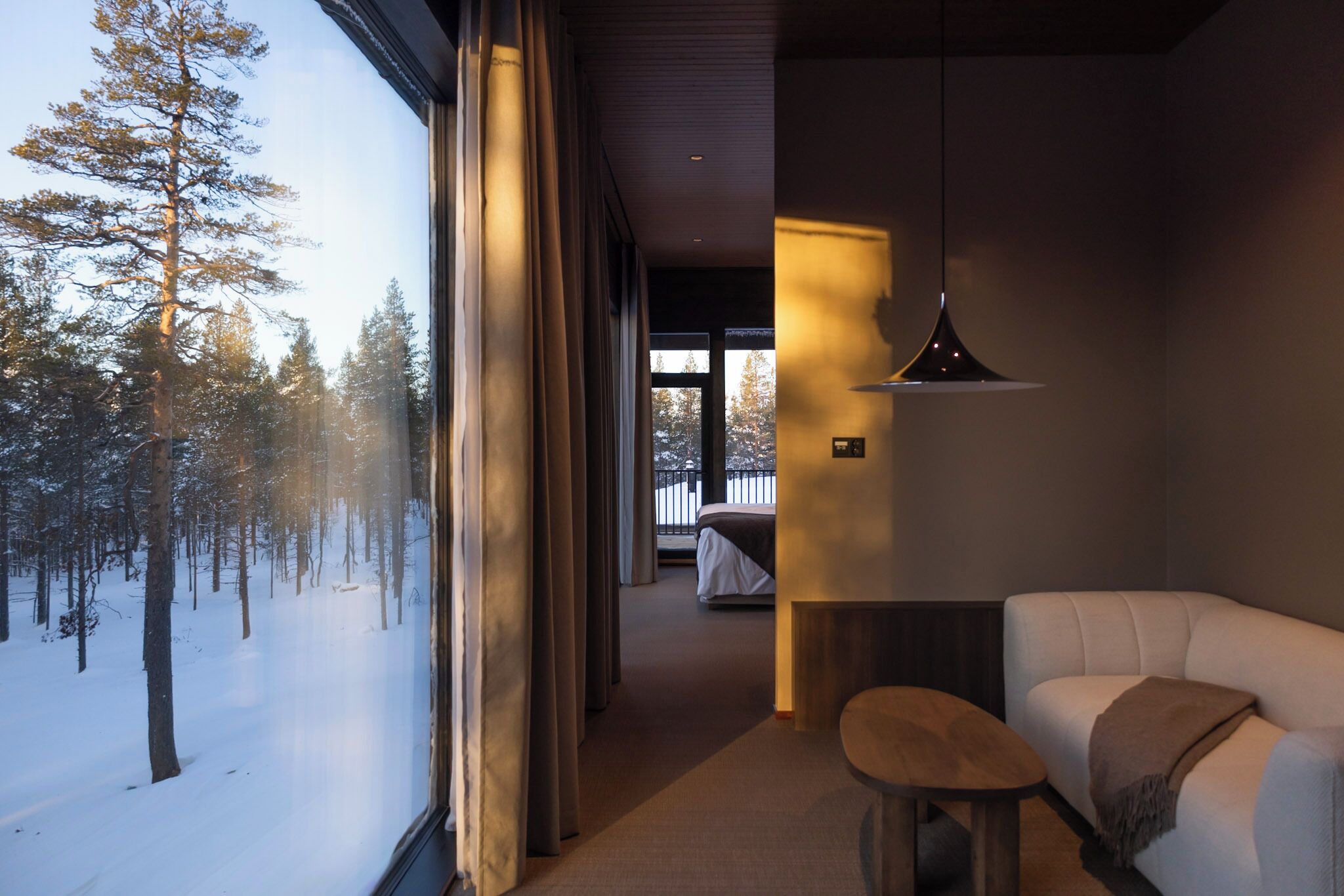 Ein modernes Hotelzimmer mit einem großen Fenster und Blick auf einen verschneiten Wald. Es gibt eine gemütliche Sitzecke mit einer Couch und einem kleinen Tisch.