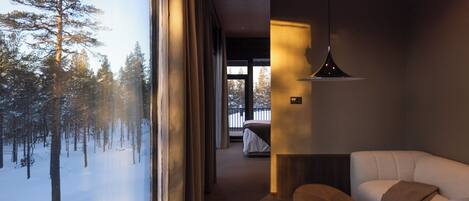 Ein modernes Hotelzimmer mit einem großen Fenster und Blick auf einen verschneiten Wald. Es gibt eine gemütliche Sitzecke mit einer Couch und einem kleinen Tisch.