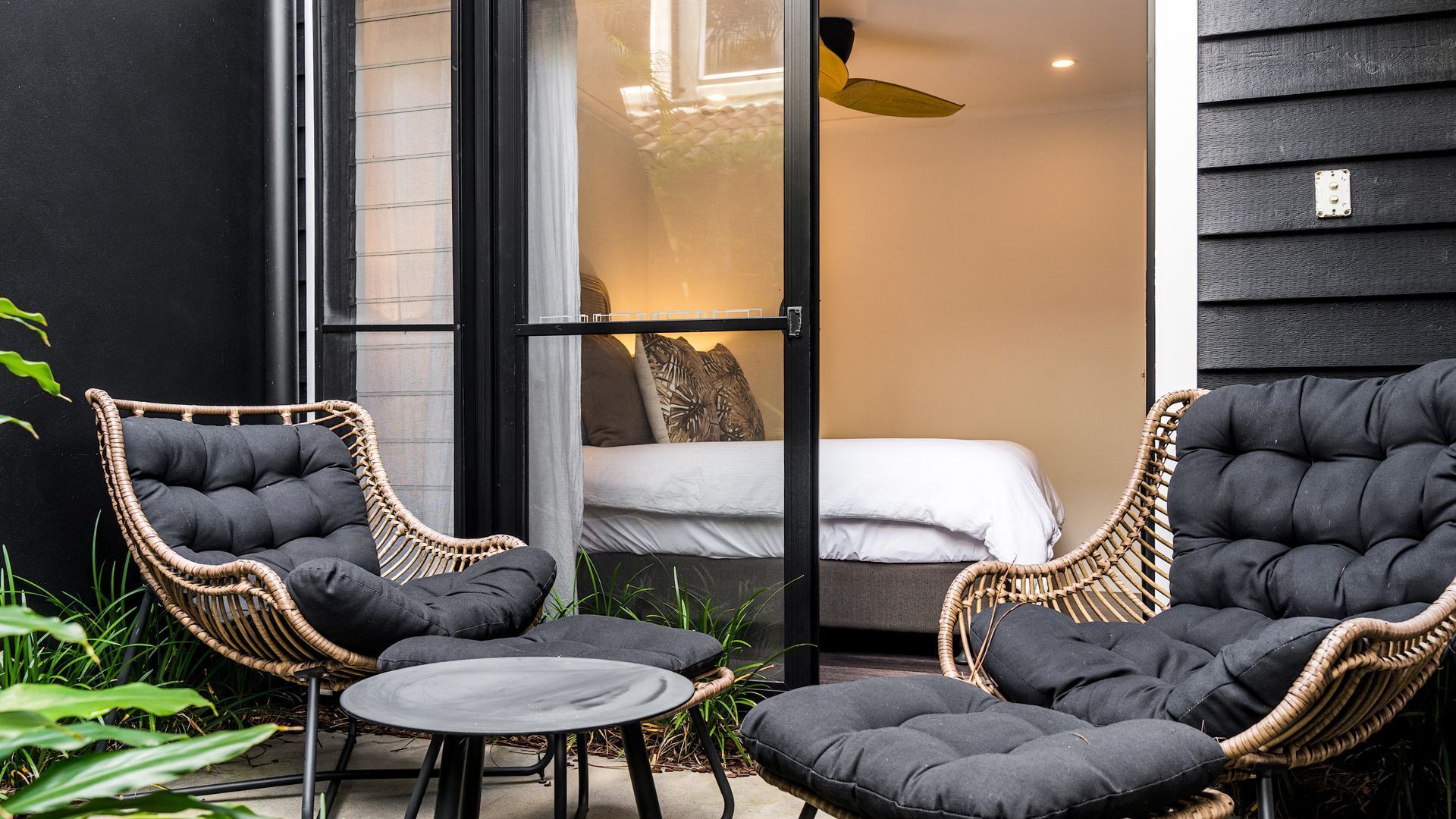 A room with a bed, two wicker chairs, a small table, and a ceiling fan.