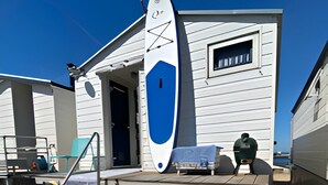 A beach house with a covered deck, a surfboard leaning against the house, and a small table with a chair. - Beach house on the North Sea coast - IJmuiden Kleine Strand (IJmuiden)