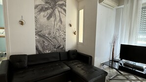 A living room with a black sectional sofa, a flat-screen TV on a glass TV stand, a wall-mounted air conditioner, and a large tropical mural. - HAN1913 (Cannes)