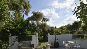 An outdoor patio with wicker furniture and a table, surrounded by lush greenery and a white wall.