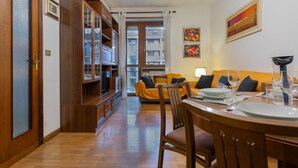 A dining area with a wooden table set for two, a sofa in the background, and a view of the outside through a glass door.
