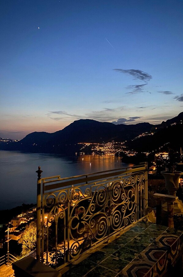 A nighttime view of a coastal town with a prominent mountain in the background, a calm sea, and a decorative metal railing in the foreground.