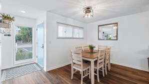 A dining area with a wooden table and chairs, a ceiling light fixture, a window with blinds, and a door leading outside. - Salt & Sun - Beach & Pool (St. Augustine)
