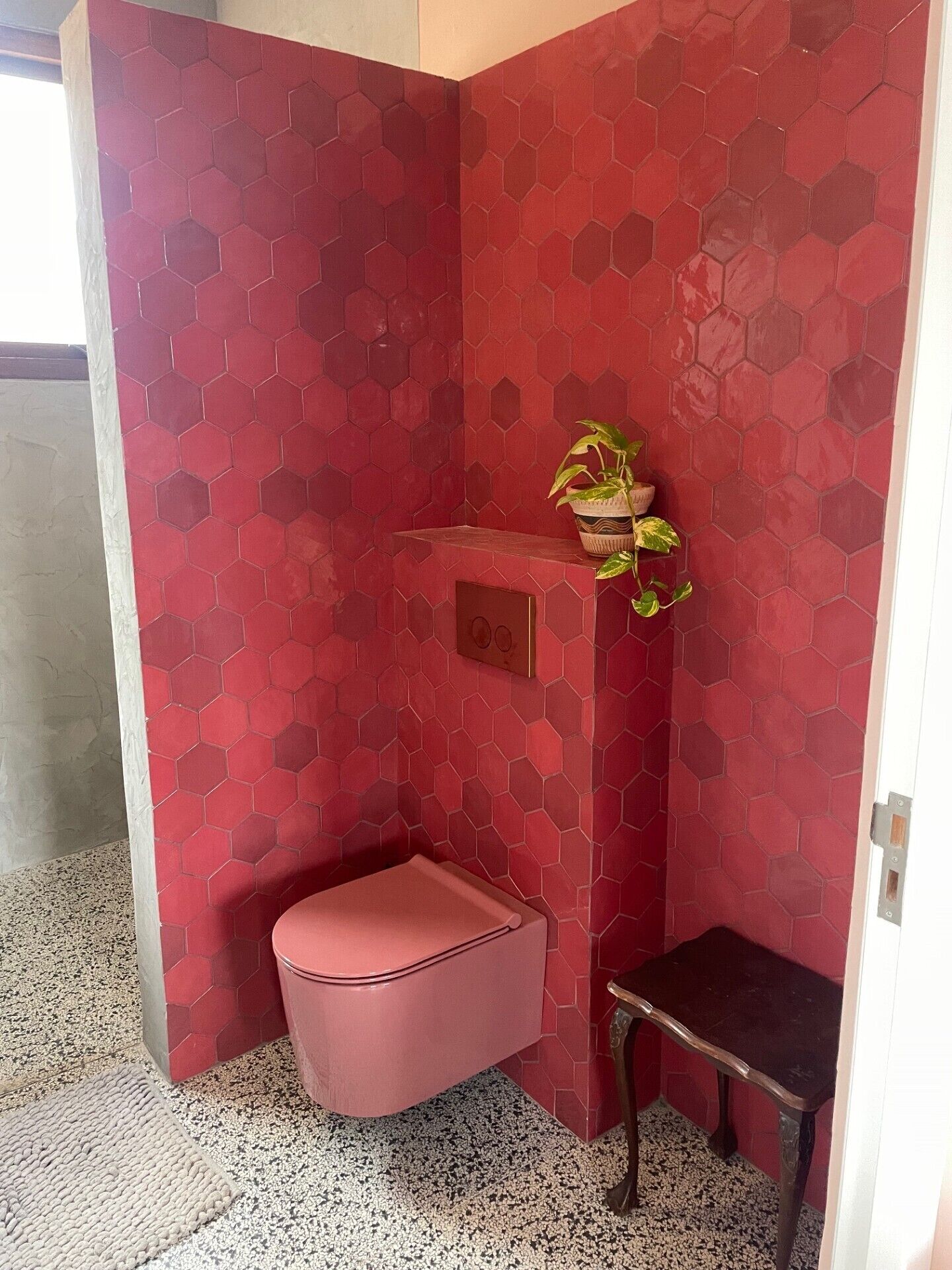 A small, red-tiled bathroom with a pink bench, a small table, and a potted plant.