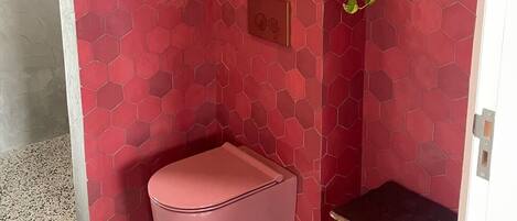 A small, red-tiled bathroom with a pink bench, a small table, and a potted plant.