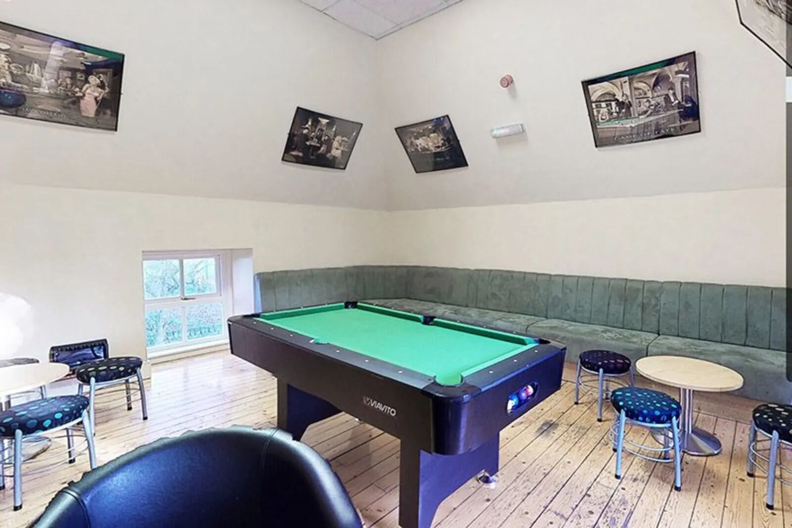 Une table de billard se trouve dans une pièce où l’on aperçoit un banc vert à coussins, une petite table et deux tabourets. Des tableaux encadrés ornent les murs.
