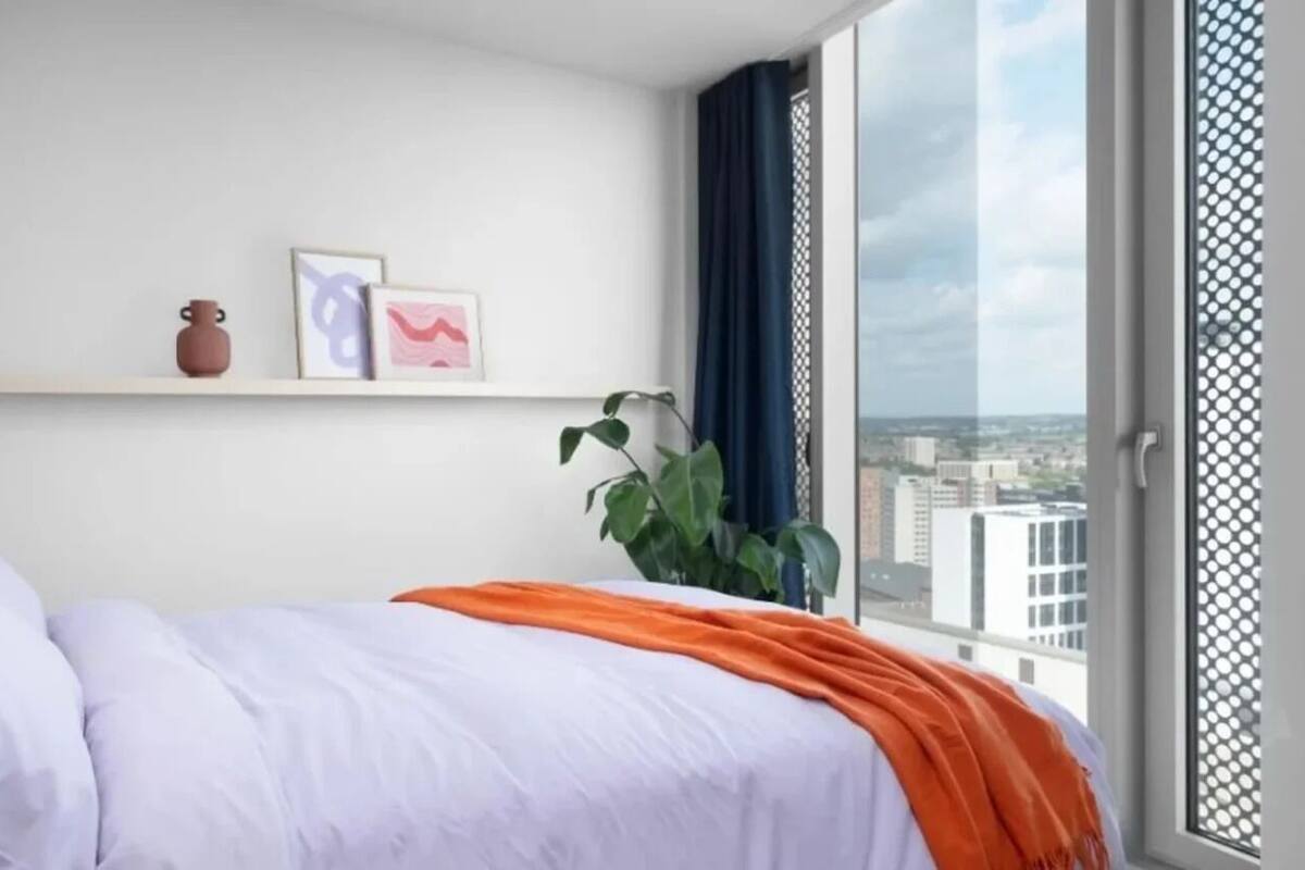 Un dormitorio con cama, ventana y vista a la ciudad, una planta y un estante con dos objetos.