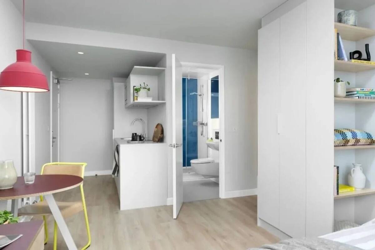 Un dormitorio moderno con cama, mesita de noche y tocador. Se observa un armario con estantes y un espejo. Se ve también un baño con ducha e inodoro.