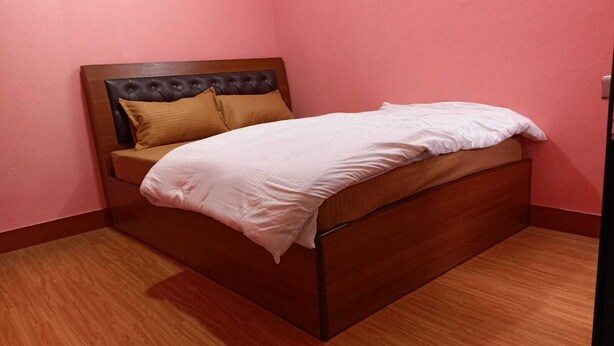 Ein Einzelbett mit Holz Kopfteil und braunem Kissen, vor einer rosafarbenen Wand mit Holzboden.