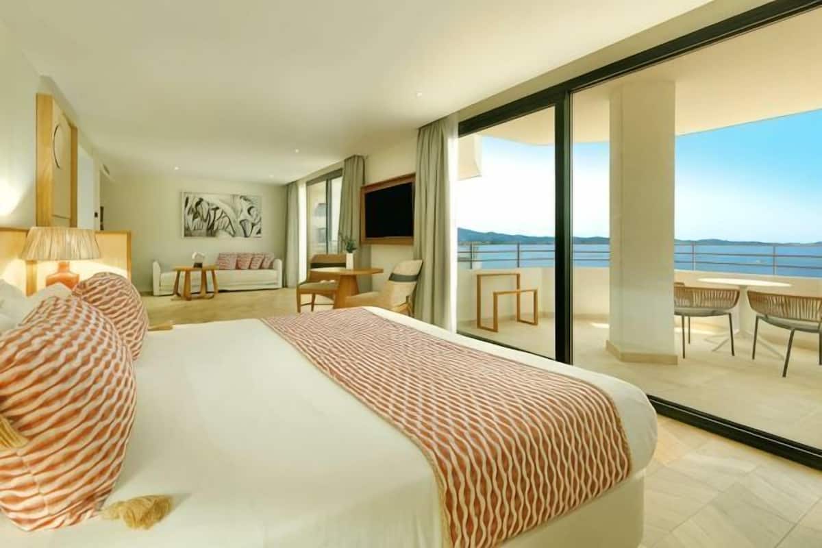 A hotel room with a large bed, a sofa, a TV, and a balcony with a view of the sea.