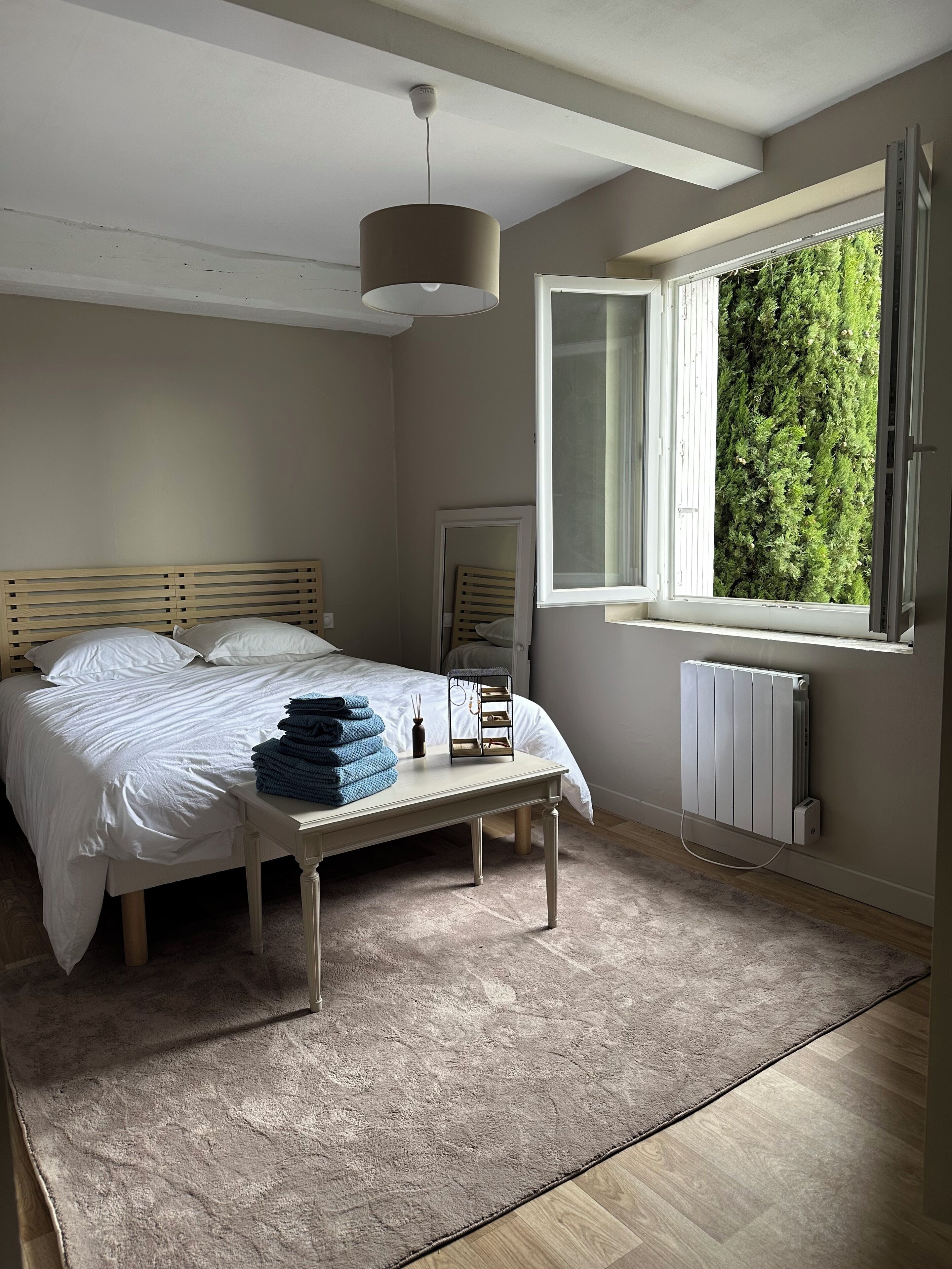 A bedroom with a bed, bedside table, lamp, window, radiator, and a view of greenery outside.
