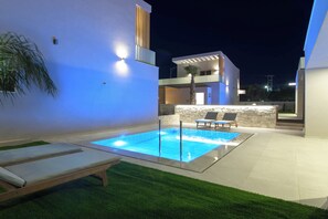 A modern outdoor pool area with a glass enclosure, two lounge chairs, and a stone wall backdrop. - Lumina Sense Villas (Kato Gouves)