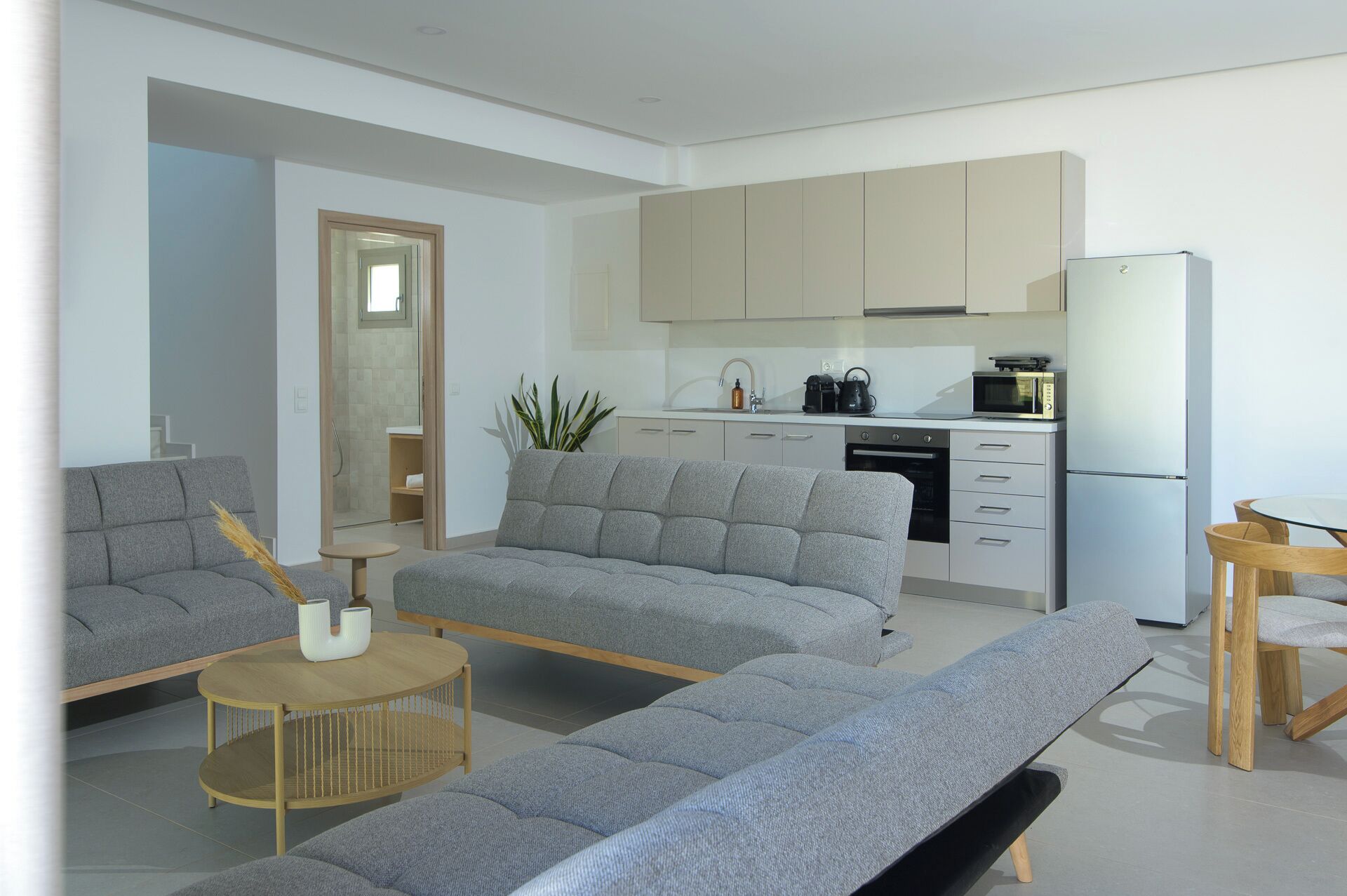 A modern living room with grey sofas, a small round table, and a kitchen area with a microwave, kettle, and cabinets.