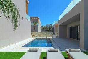 A modern outdoor pool area with a stone wall, a palm tree, and lounge chairs with towels. - Lumina Sense Villas (Kato Gouves)