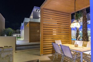 An outdoor patio area with a wooden slatted wall, a table with chairs, and a hanging light fixture. - Lumina Sense Villas (Kato Gouves)