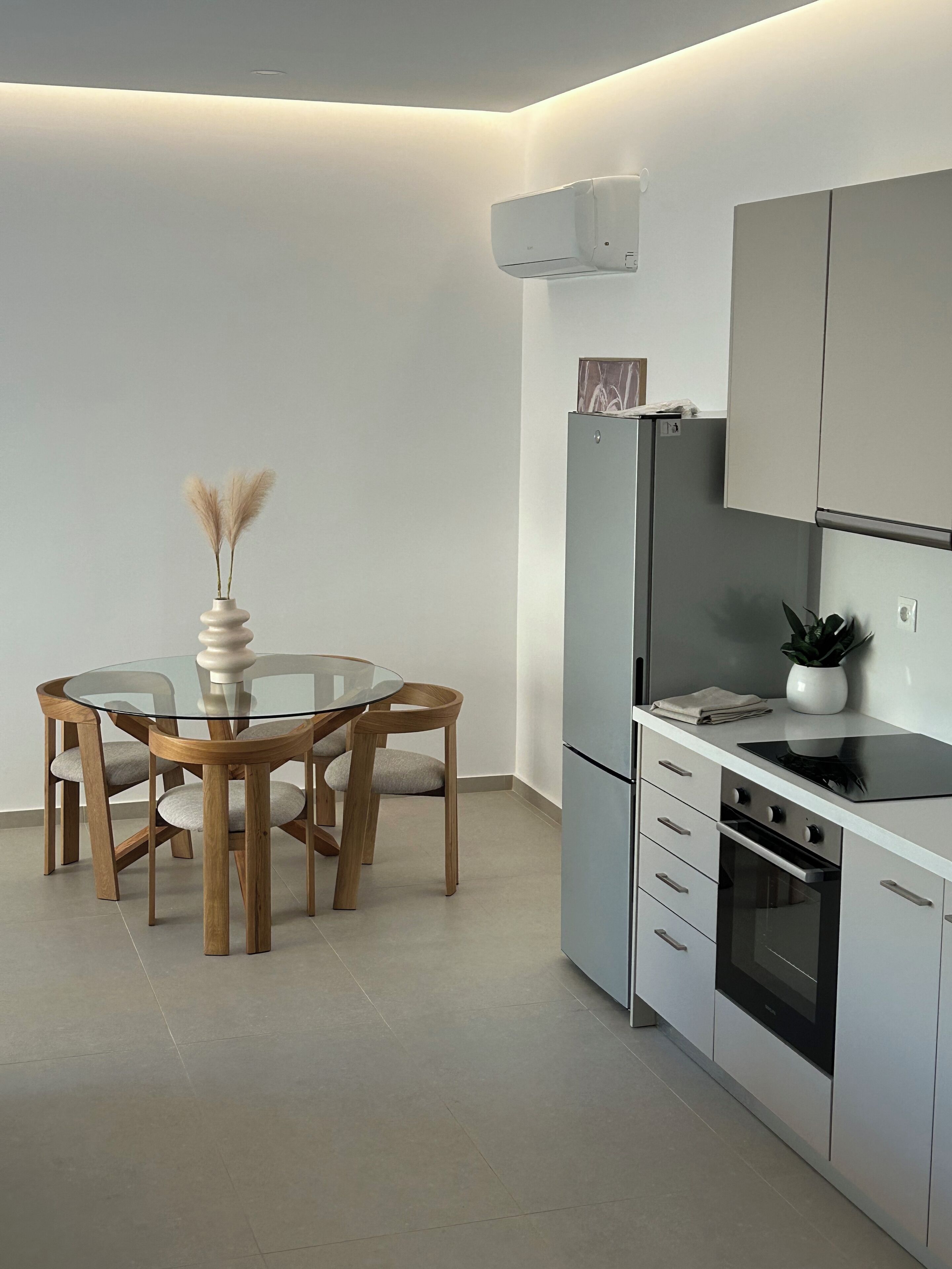 A modern kitchen with a round glass table, wooden chairs, a built-in oven, and a grey cabinet.