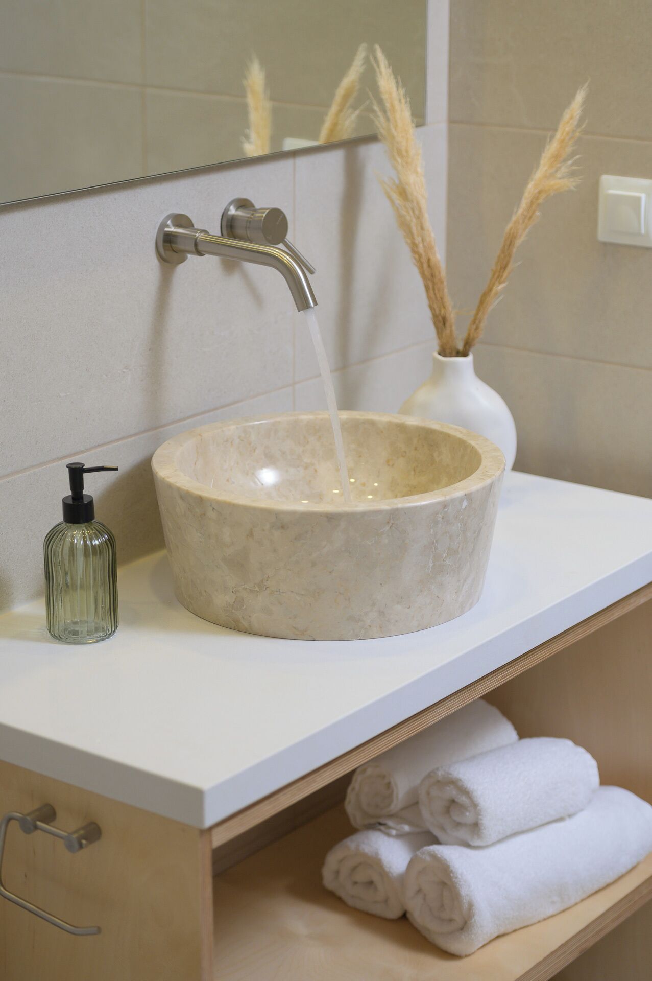 A bathroom with a stone sink, a faucet, a soap dispenser, a vase with pampas grass, and a towel rack with towels.