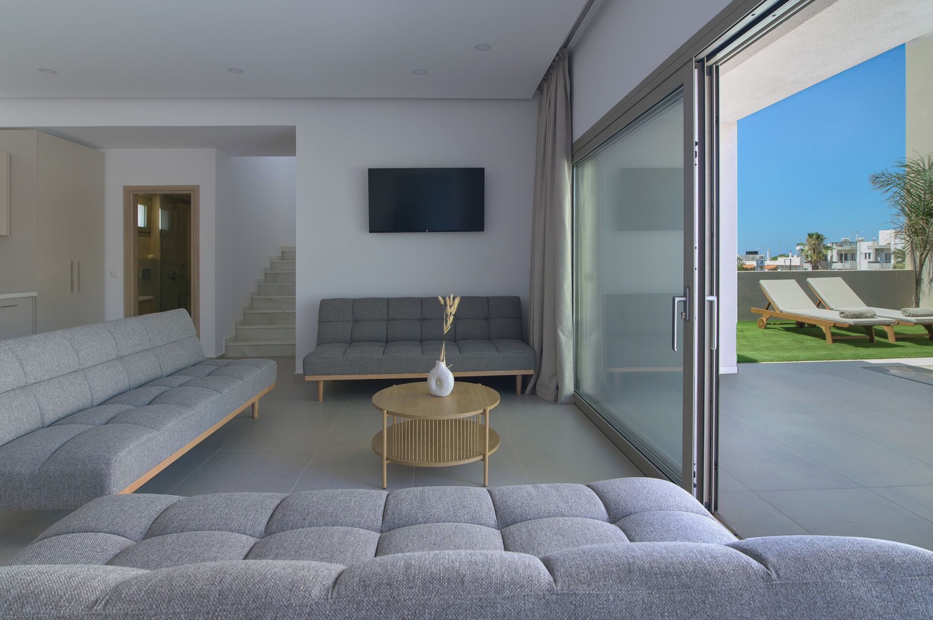 Un saló modern amb un sofà gris, un petit divà gris i una taula de centre rodona amb un vas. Una televisió de pantalla plana està penjada a la paret, i una porta corredissa de vidre dona pas a una zona exterior amb gandules.