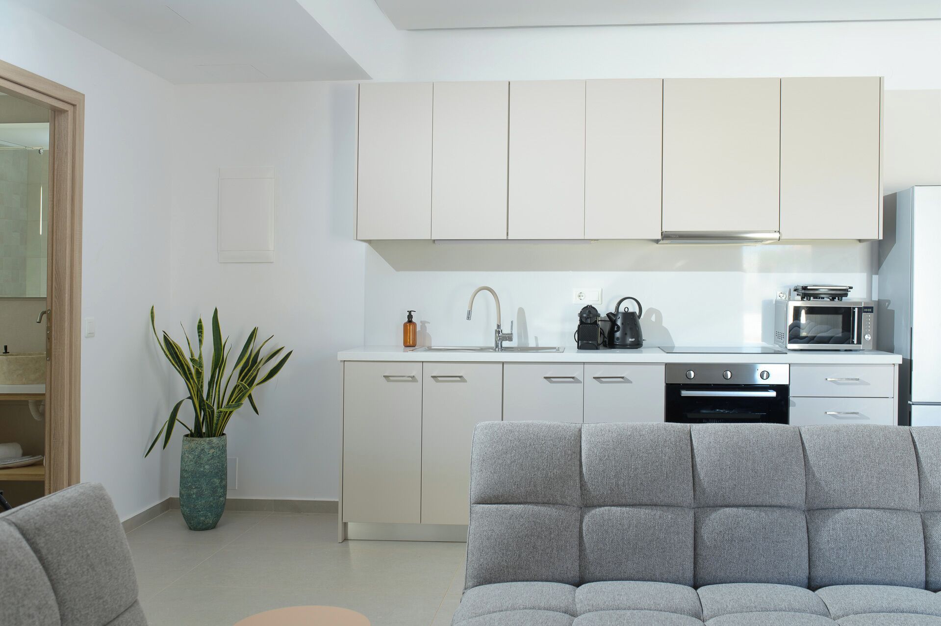 A modern kitchen with white cabinets, a built-in oven, and a microwave. There is a grey sofa in the foreground and a potted plant to the left.