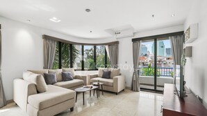 A modern living room with a sectional sofa, a coffee table, and a TV on a wooden cabinet. Large windows offer a view of the cityscape. - Beleria by Kozystay - Senayan (Jakarta)