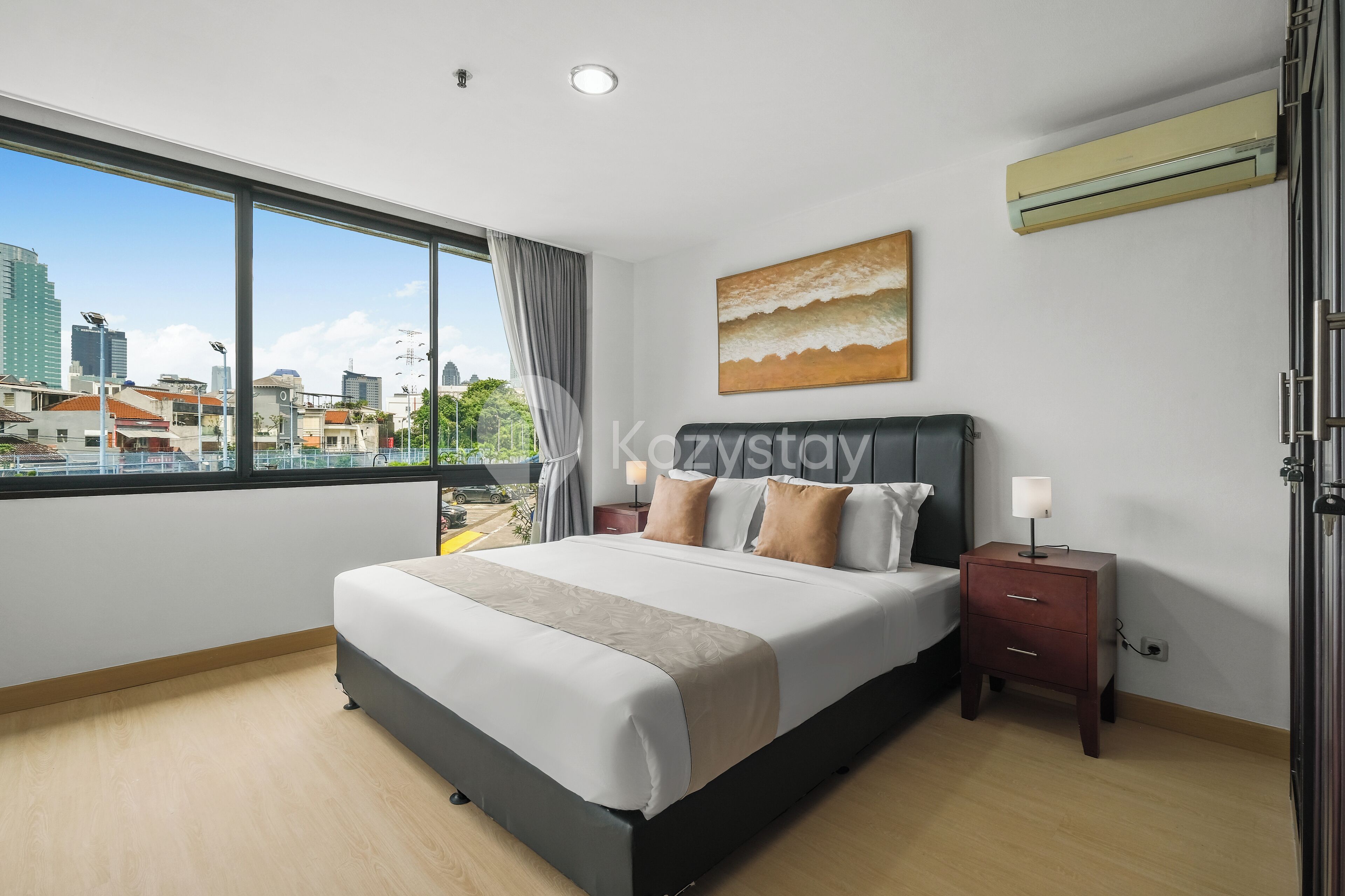 A modern hotel room with a large bed, a nightstand, a bedside lamp, and a city view through large windows.