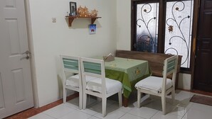 A dining area with a table and four chairs, a shelf with decorative items, a door, and a window with decorative ironwork. - Ventera Homestay (Kalasan)